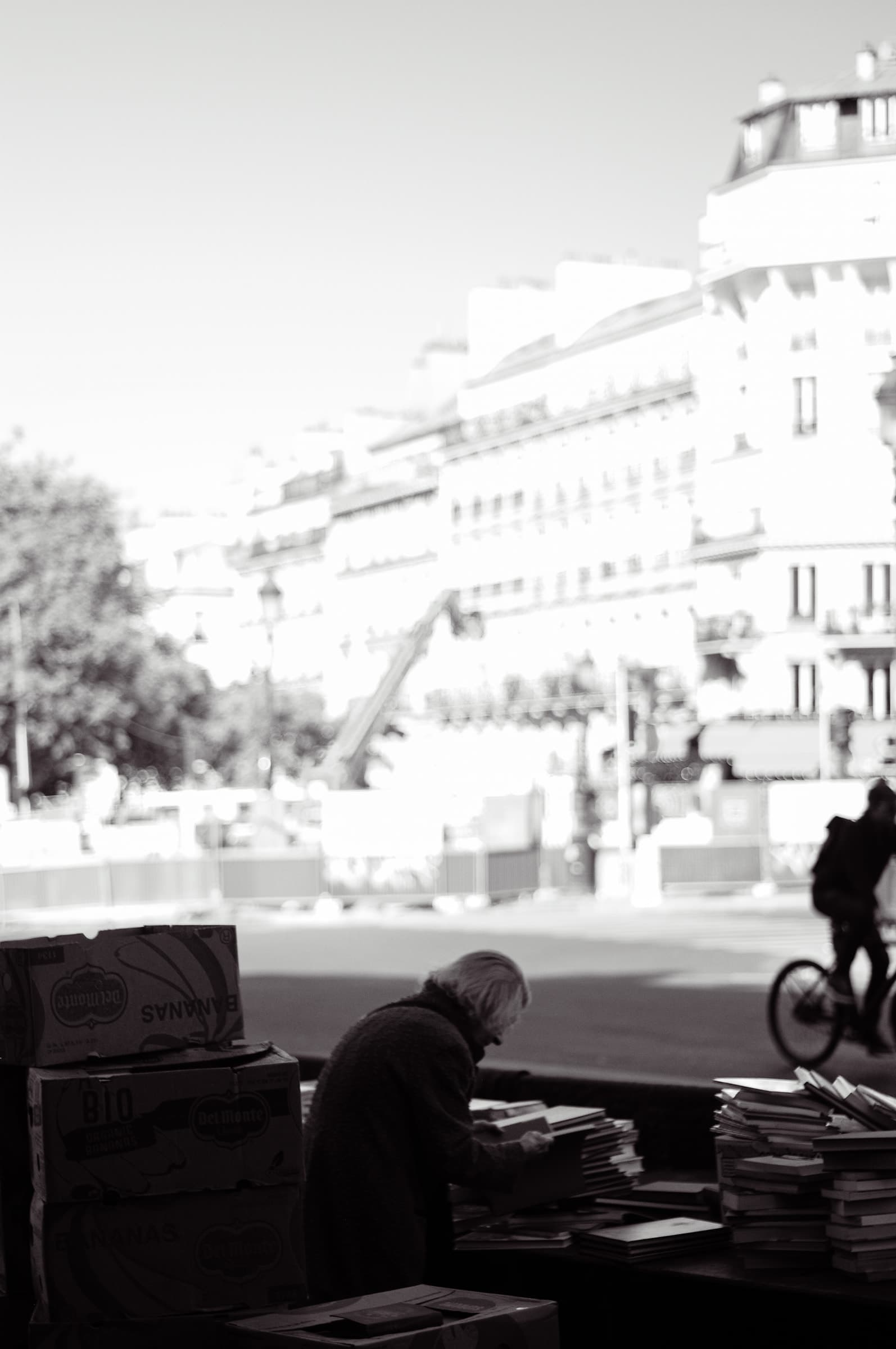 A person seated beside stacks of books in the shade at the edge of the square