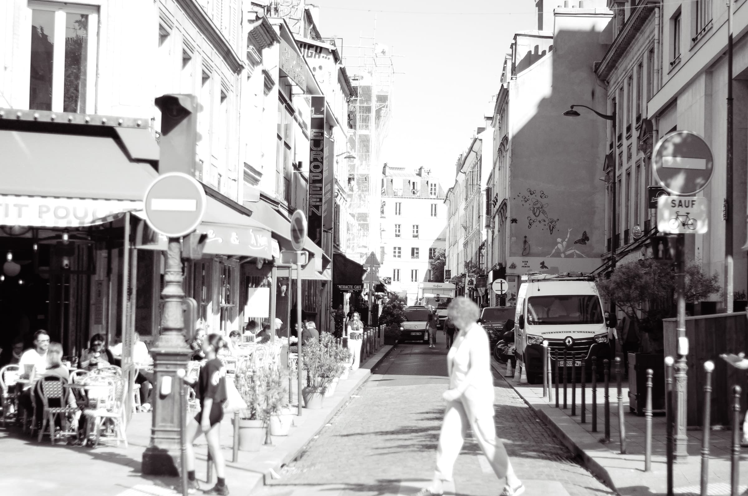 A person crossing a narrow side street lined with cafés and parked cars